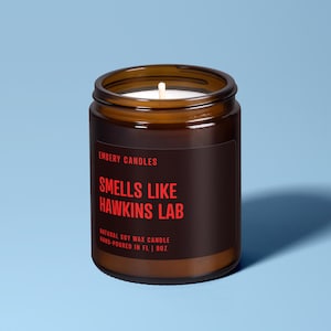 May include: A brown glass jar candle with a black label featuring red text that reads EMBERY CANDLES, SMELLS LIKE HAWKINS LAB, NATURAL SOY WAX CANDLE, HAND-POURED IN FL | 9OZ. The candle has a white wick centered in the wax inside the glass container.