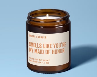 Könnte beinhalten: Eine Kerze im braunen Glasgefäß mit einem weißen Etikett, das den Text EMBERY CANDLES, SMELLS LIKE YOU'RE MY MAID OF HONOR, NATURAL SOY WAX CANDLE, HAND-POURED IN FL | 9OZ trägt. Die Kerze hat einen sichtbaren weißen Docht im Wachs.