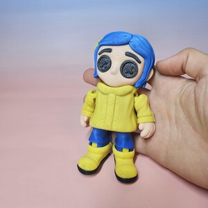 May include: A 3D-printed figurine of a character with blue hair, button eyes, and a yellow coat. The figure is wearing blue pants and yellow boots. The figurine is held in a hand against a pink background.
