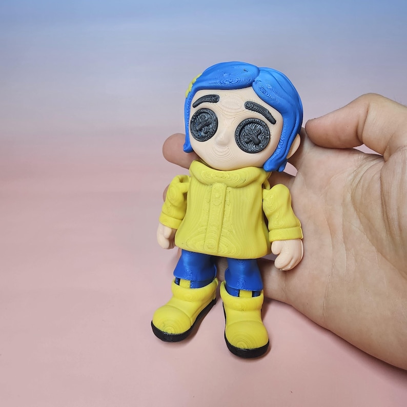May include: A 3D-printed figurine of a character with blue hair, button eyes, and a yellow coat. The figure is wearing blue pants and yellow boots. The figurine is held in a hand against a pink background.