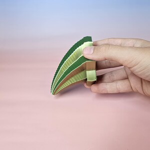 May include: A hand holding a fan-shaped object composed of layered paper in shades of green and brown. The object has a curved shape, with the colors transitioning from light to dark. The background is a gradient of pink and blue.