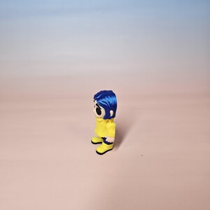 May include: A figurine of a character with blue hair, wearing a yellow jacket and yellow boots. The figurine has a stylized design with large black eyes and a neutral expression. The background is a gradient of pink and blue.