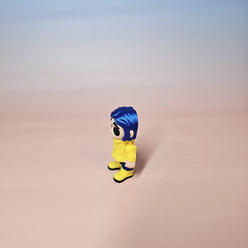 May include: A figurine of a character with blue hair, wearing a yellow jacket and yellow boots. The figurine has a stylized design with large black eyes and a neutral expression. The background is a gradient of pink and blue.