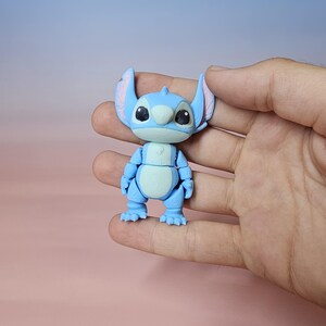 May include: A light blue articulated Stitch figure with large ears, black eyes, and a white belly. The figure is held in a hand against a soft pink and blue background. The toy is a collectible item.