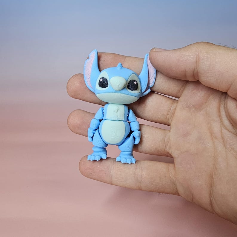 May include: A light blue articulated Stitch figure with large ears, black eyes, and a white belly. The figure is held in a hand against a soft pink and blue background. The toy is a collectible item.