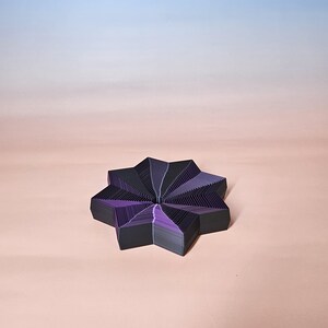 May include: A geometric, star-shaped object with a layered design. The object features alternating black and purple sections, creating a visually striking pattern. The background is a gradient of pink and blue.
