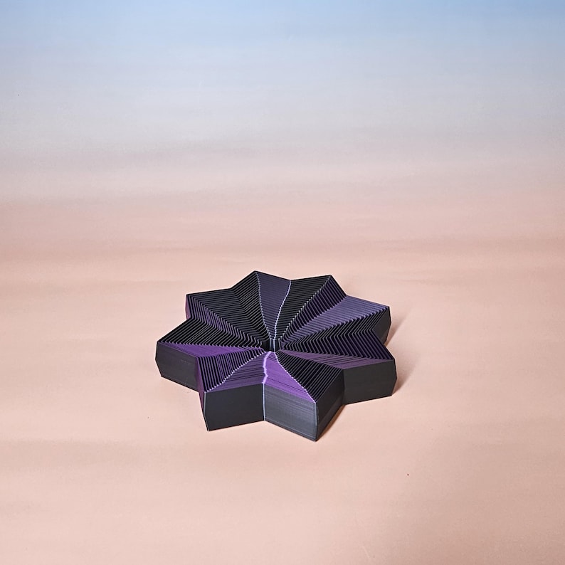 May include: A geometric, star-shaped object with a layered design. The object features alternating black and purple sections, creating a visually striking pattern. The background is a gradient of pink and blue.