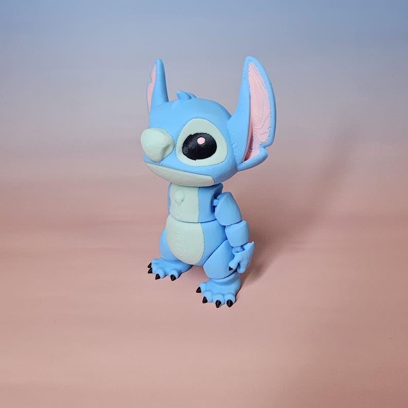 May include: A blue articulated figure of a cartoon character with large ears, a large eye, and a white nose. The figure has a light blue body with darker blue accents and black claws. The background is a gradient of pink and blue.