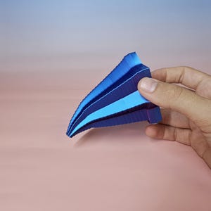 May include: A geometric, abstract sculpture in shades of blue, held by a hand. The sculpture has a layered, fan-like design with varying tones of blue, from deep navy to light sky blue. The object is set against a gradient background.