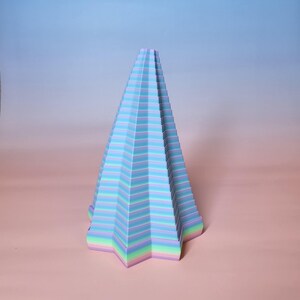 May include: A pastel-colored, geometric, cone-shaped sculpture. The sculpture features horizontal stripes of blue, purple, and green. The base is a five-pointed star. The artwork is set against a gradient background of pink and blue.