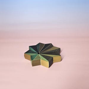May include: A star-shaped decorative object with a layered design. The object features a combination of green, gold, and black colors. The background is a gradient of pink and blue.