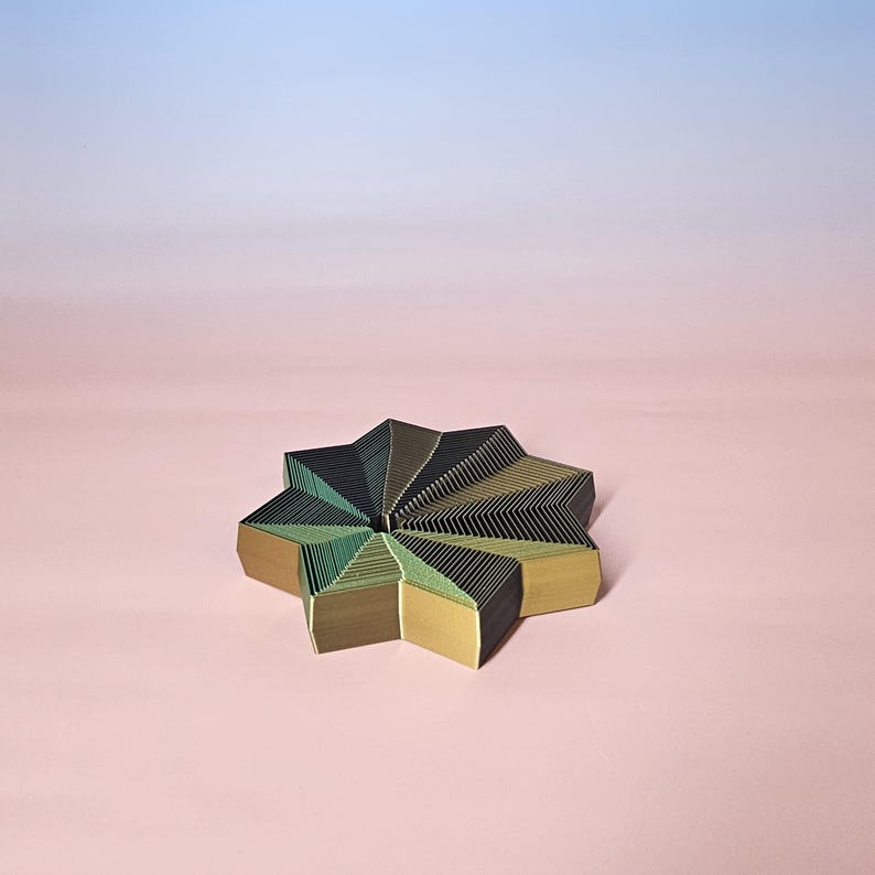 May include: A star-shaped decorative object with a layered design. The object features a combination of green, gold, and black colors. The background is a gradient of pink and blue.