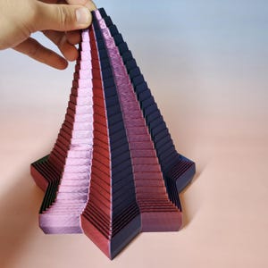May include: A multi-layered, star-shaped sculpture with a tiered design. The sculpture features alternating bands of red, pink, and dark blue. The object is held by a hand, showcasing its intricate, geometric form. The sculpture has a unique, artistic appearance.