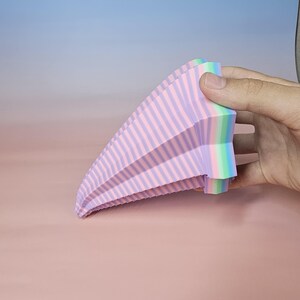 May include: A multi-colored, fan-shaped object with pink and purple stripes. The object is held by a hand and has a rainbow-colored edge. The background is a gradient of pink and blue.