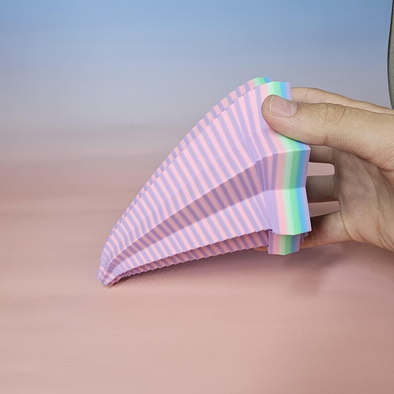 May include: A multi-colored, fan-shaped object with pink and purple stripes. The object is held by a hand and has a rainbow-colored edge. The background is a gradient of pink and blue.