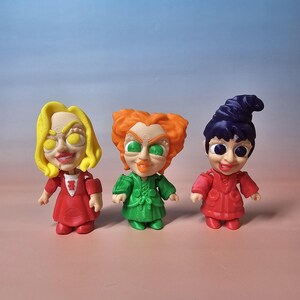 May include: Three colorful figurines of the Sanderson sisters from the movie Hocus Pocus. Each figure has a unique hairstyle and outfit. The first is yellow-haired in a red dress, the second has orange hair and a green dress, and the third has a purple hat and a red dress.