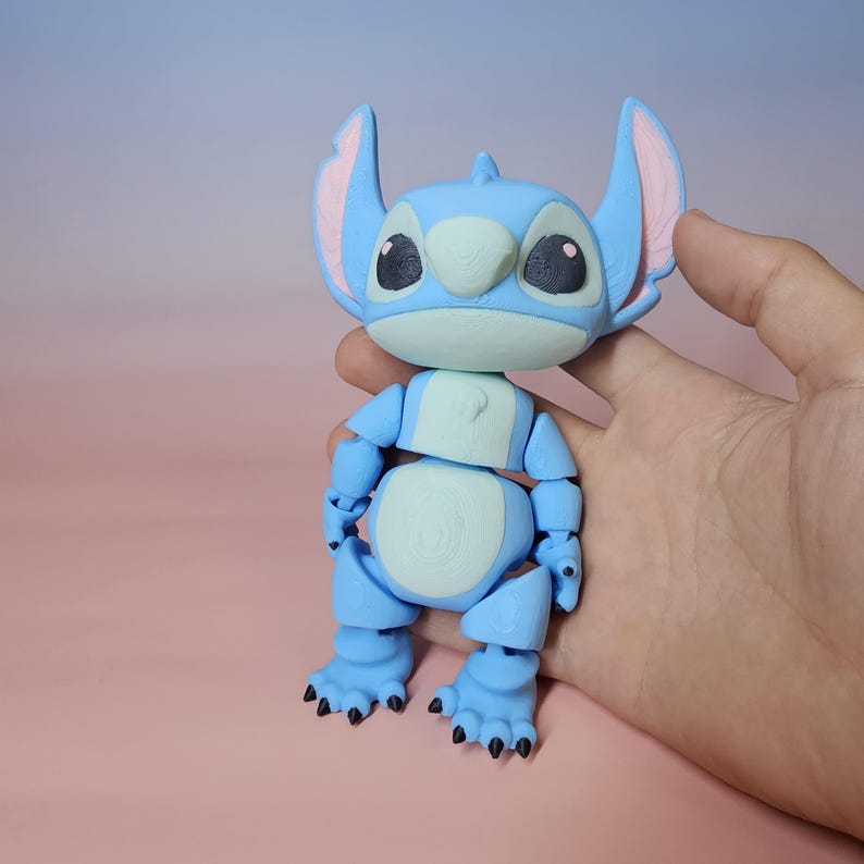 May include: A light blue articulated Stitch figure, featuring large ears with pink inner details, black eyes, and a light green belly. The figure is from the animated film Lilo & Stitch and is held in a hand.
