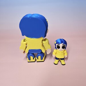 May include: Two figurines of a character from a movie. The larger figurine is yellow and blue, with a yellow jacket and blue hair. The smaller figurine is also yellow and blue, with a yellow sweater and blue hair. The figurines are on a pink surface.