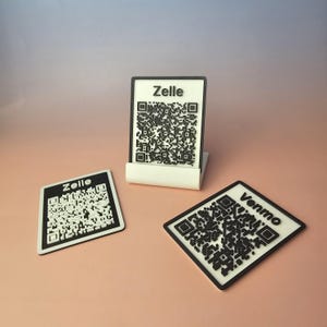 May include: Three payment QR code displays. One upright in a white stand, and two flat. The displays have black and white QR codes with the words "Zelle" and "Venmo" printed on them. The background is a solid light pink.
