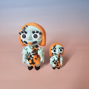 May include: Two handmade figurines of a character with a light blue face, orange hair, and a patchwork dress. The larger figure is approximately 10 cm tall, and the smaller one is about 5 cm tall. The figures have black accents and large, round eyes.