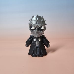 May include: A silver and black figurine of a character with a spiked head and a black robe. The figure has a detailed face with large eyes and a stern expression. The figurine is standing on a solid surface.