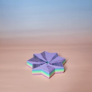 May include: A star-shaped object composed of layered, light purple paper with a striped pattern. The edges of the star display a rainbow gradient of colors. The background is a gradient of light blue and peach.