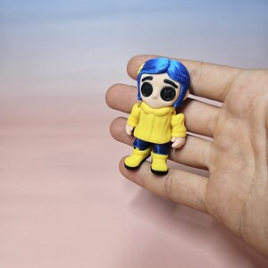 May include: A small figurine of a character with blue hair, wearing a yellow jacket, blue pants, and yellow boots. The figurine has large black eyes and a neutral expression. The figure is held in a hand against a pink and blue background.
