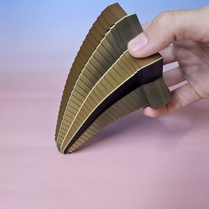 May include: A fan-shaped collection of brown and gold samples, held in a hand against a pink and blue gradient background. The samples have a layered design with a curved shape, showcasing a range of shades and textures. The edges are clean and precise.