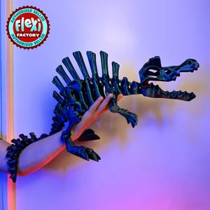 Puede incluir: Un modelo de esqueleto de dinosaurio de plástico negro con una superficie texturizada. El dinosaurio está en posición con la boca abierta y la cabeza inclinada hacia un lado. El dinosaurio está siendo sostenido por la mano de una persona.