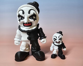 May include: Two clown figurines, one large and one small, stand side-by-side. The figures are split vertically, with one half white and the other black. They have white faces with black eye makeup, red mouths, and small black hats.