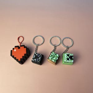 May include: Four pixelated keychains. One is a red and black heart shape. The other three are square-shaped, with pixelated designs in black and white, green and brown, and green and black. Each keychain has a silver ring.