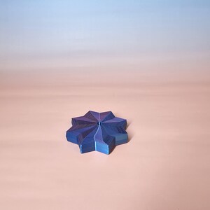 May include: A blue and purple star-shaped object with a textured surface. The object is made of a series of layered, angled planes. The background is a gradient of pink and blue.