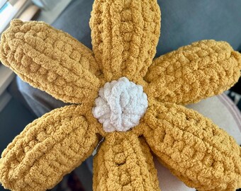 May include: A crocheted flower pillow in a mustard yellow color with a white center. The pillow is made with a fluffy yarn and has a soft, plush texture.