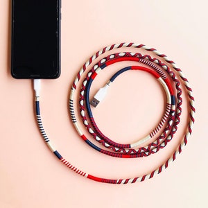 Può includere: Uno smartphone nero con un cavo di ricarica bianco avvolto in tessuto rosso, bianco e blu. Il cavo è avvolto e collegato al telefono.