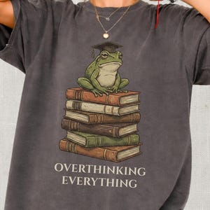 May include: Dark gray t-shirt featuring a green frog wearing a graduation cap, sitting atop a stack of books. The text "OVERTHINKING EVERYTHING" is printed below the image in white. The shirt has short sleeves and a relaxed fit.