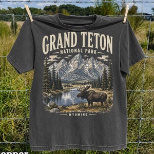 May include: A charcoal gray t-shirt featuring a graphic print of the Grand Teton National Park in Wyoming. The design includes a detailed illustration of snow-capped mountains, a pine forest, a lake, and a moose, with the text GRAND TETON NATIONAL PARK.
