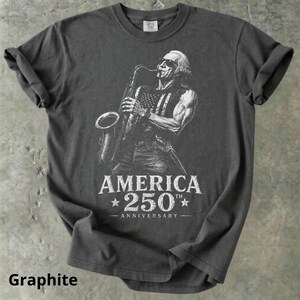 May include: Graphite-colored t-shirt featuring a white illustration of a person playing a saxophone with an American flag design on their vest. The text "AMERICA 250th ANNIVERSARY" is below the illustration. The shirt has rolled-up sleeves.