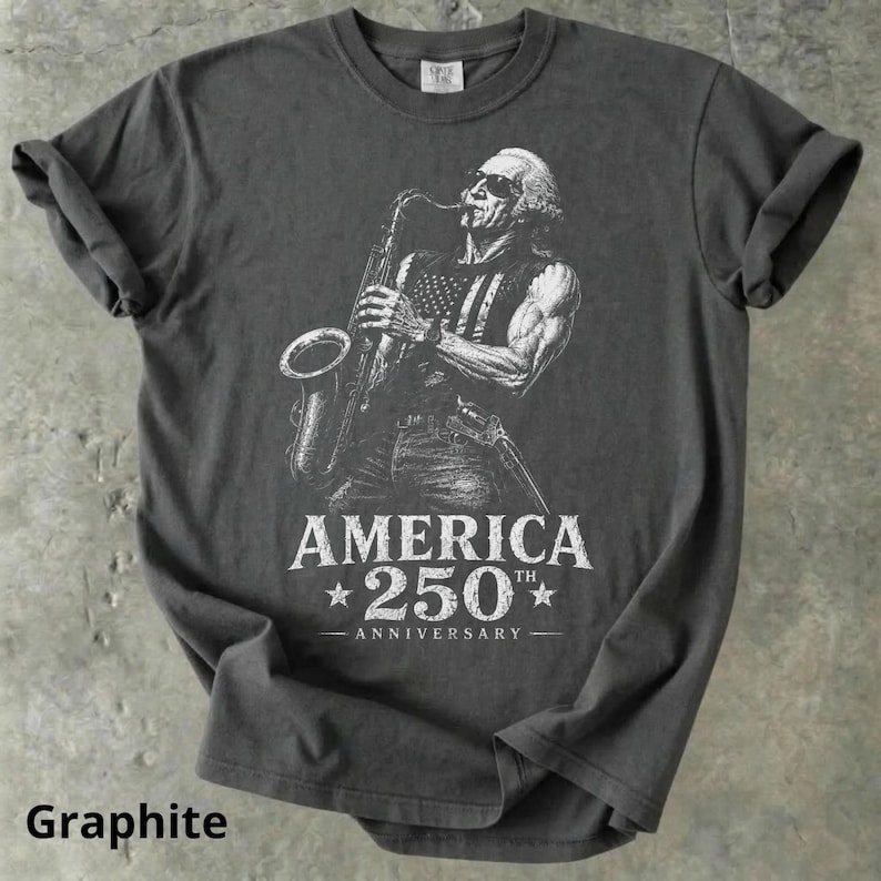 May include: Graphite-colored t-shirt featuring a white illustration of a person playing a saxophone with an American flag design on their vest. The text "AMERICA 250th ANNIVERSARY" is below the illustration. The shirt has rolled-up sleeves.