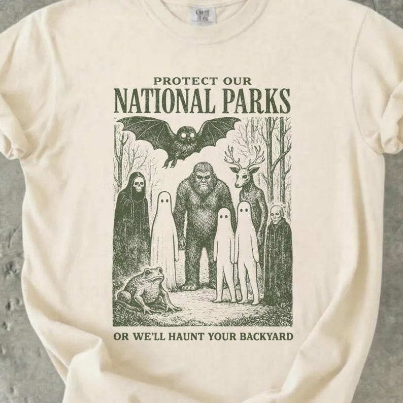 Ivory Comfort Colors 1717 shirt with vintage dark green cryptid illustration featuring Mothman, Bigfoot, ghosts, a deer, and a frog, with the text “Protect Our National Parks Or We’ll Haunt Your Backyard.”