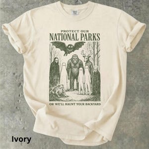 May include: Ivory-colored t-shirt with a graphic print. The design features a bat, Bigfoot, a deer, ghosts, a frog, and the text "PROTECT OUR NATIONAL PARKS OR WE'LL HAUNT YOUR BACKYARD."