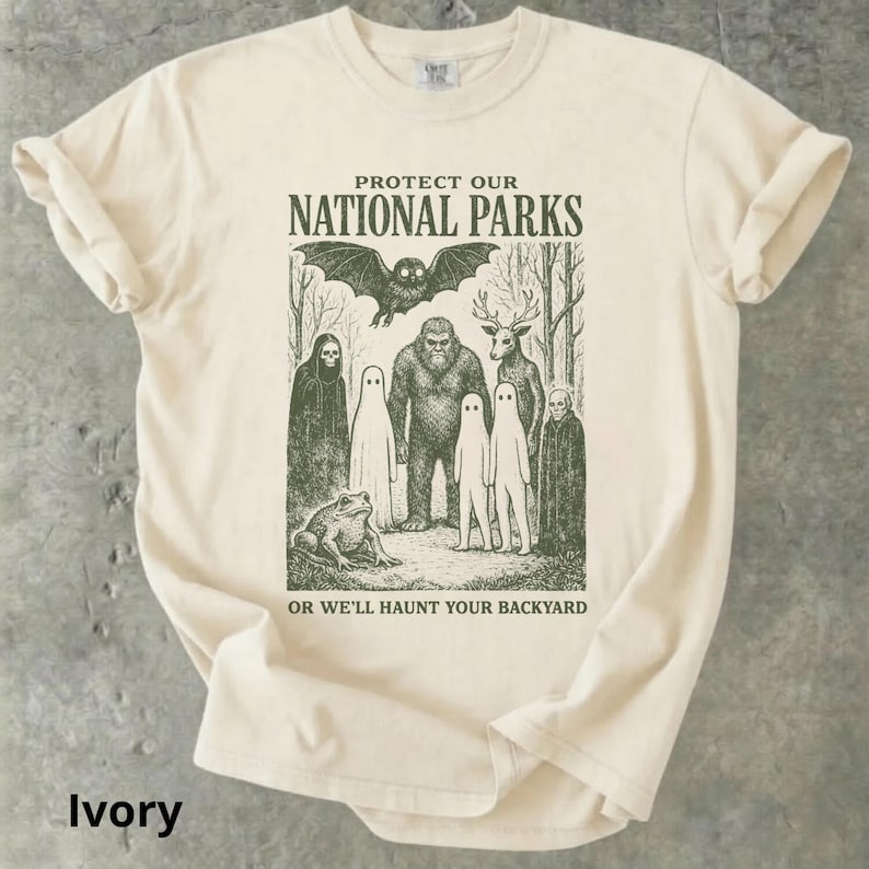 May include: Ivory-colored t-shirt with a graphic print. The design features a bat, Bigfoot, a deer, ghosts, a frog, and the text "PROTECT OUR NATIONAL PARKS OR WE'LL HAUNT YOUR BACKYARD."