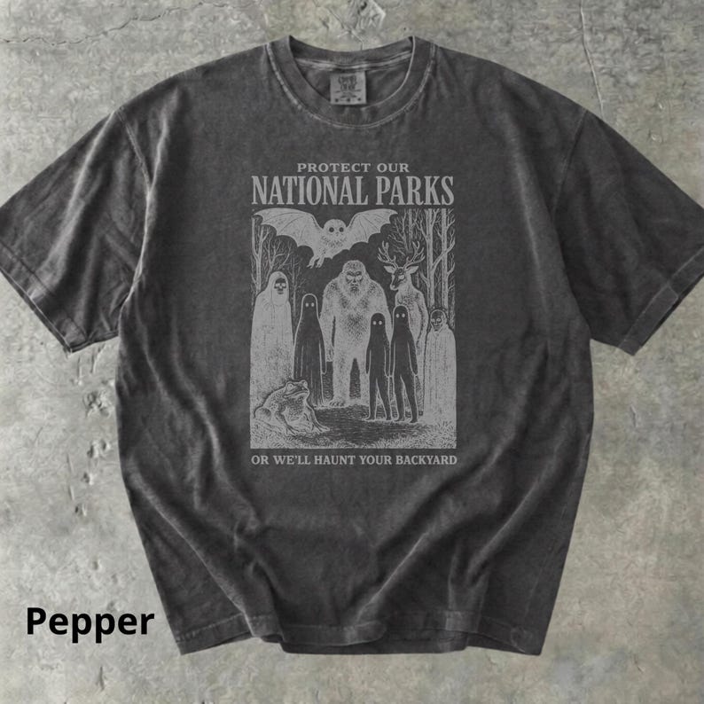 May include: Pepper-colored t-shirt with a graphic that reads "PROTECT OUR NATIONAL PARKS" above an illustration of forest creatures. Below the graphic, the text reads "OR WE'LL HAUNT YOUR BACKYARD".