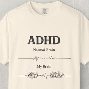 May include: A cream-coloured t-shirt featuring the word "ADHD" in dark, bold letters. Underneath, it reads "Normal Brain" with a straight line, and "My Brain" with a scribbled line. The shirt is made of a soft material.