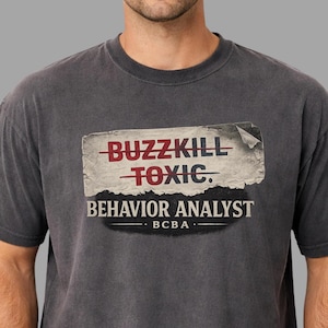May include: Dark grey t-shirt featuring the text "BUZZKILL" crossed out and replaced with "TOXIC" in red, above "BEHAVIOR ANALYST" and "BCBA" in white. The shirt has a vintage, distressed appearance.