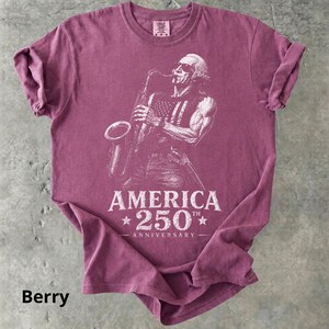 May include: Berry-colored t-shirt featuring a detailed illustration of a musician playing a saxophone, with an American flag design on the vest. The text "AMERICA 250th ANNIVERSARY" is printed below the image.