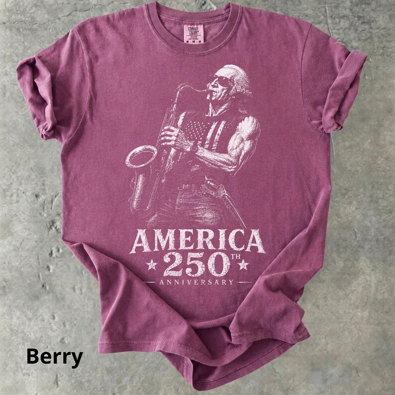 May include: Berry-colored t-shirt featuring a detailed illustration of a musician playing a saxophone, with an American flag design on the vest. The text "AMERICA 250th ANNIVERSARY" is printed below the image.