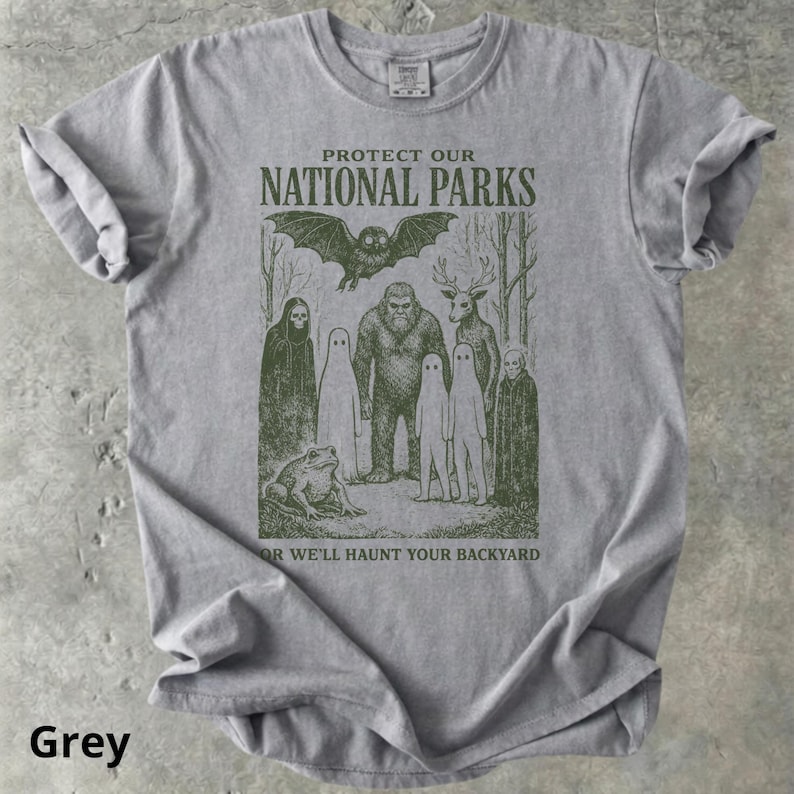May include: Grey t-shirt with a graphic featuring a bat, Bigfoot, deer, ghosts, and a frog. The text reads "PROTECT OUR NATIONAL PARKS" above the illustration and "OR WE'LL HAUNT YOUR BACKYARD" below. The shirt is a comfortable, casual style.