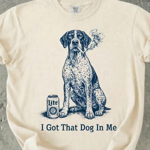 Pode incluir: Camiseta creme com uma ilustração de um cão sentado fumando um cigarro. Uma lata de cerveja está ao lado do cão. O texto "I Got That Dog In Me" está impresso abaixo. O cão tem uma pelagem manchada.