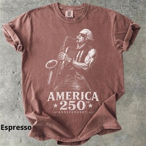 May include: Espresso-colored t-shirt featuring a white graphic of a musician playing a saxophone, with an American flag design on the vest. The text "AMERICA 250th ANNIVERSARY" is printed below the image.