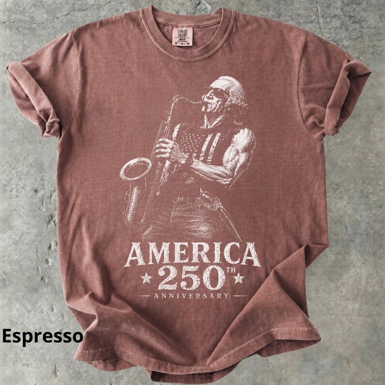 May include: Espresso-colored t-shirt featuring a white graphic of a musician playing a saxophone, with an American flag design on the vest. The text "AMERICA 250th ANNIVERSARY" is printed below the image.